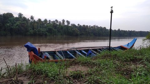 Small old blue fishing boat tied to a light pole near riverside with green coconut trees background during kerala heavy wind and rain flood because of fast moving or flowing dirty brown river water.