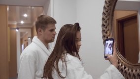 A young couple in white terry bathrobes in a spa salon shoot a video at the mirror. Vacation
