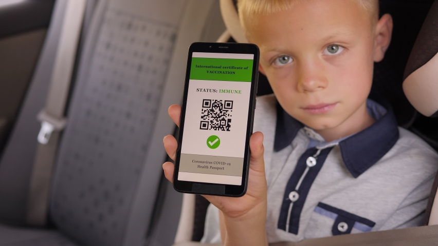 A little boy is sitting in a car fastened with seat belts and shows a QR code on the phone about vaccination against coronavirus infection.