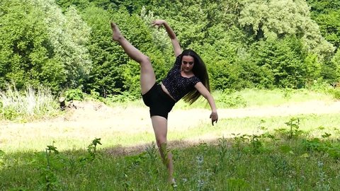 A sports girl is demonstrating her flexible legs, performing splits while standing barefoot on prickly grass in slow motion. Gymnastic  Workout Outside. Flexibility Exercises. Stretching  Outdoor.