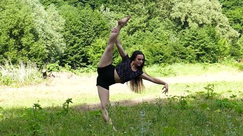 Young girl standing raises her leg and demonstrates leg stretching outdoors, slow motion. Gymnast doing splits while standing in the forest in summer. Outdoor Flexibility, Stretching Exercises. 