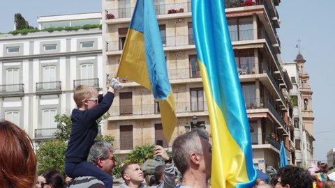 VALENCIA, SPAIN - MARCH 27, 2022: Protest against war in Ukraine. Protest against Russia's invasion of Ukraine. People gathered in the city square, holding Ukrainian flags in their hands. The boy sits