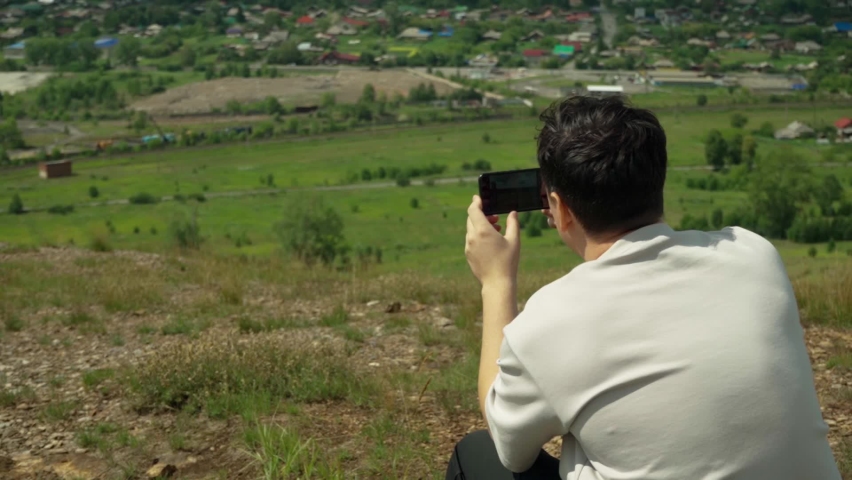 A young adult male takes a smartphone picture from the top of a large hill. Pan