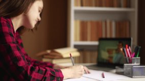 learn virtual. a teenager girl is studying remotely from a teacher online on a laptop sitting at a table next to a bookcase. indoor distance learning education concept. student on exam online