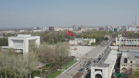 Bushkek, Kyrgyzstan - April 3, 2020: Aerial cinematic footage of red Kyrgyz national flag winding in wind in golden sun light against panoramic view of centre of Bishkek. Kyrgyzstan symbol in city. 