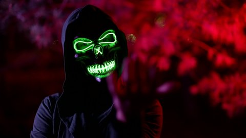 portrait of a person in a terrible glowing mask of death and in a hood against a background of branches illuminated by red light. the character beckons to him, then curls fingers. the dark key