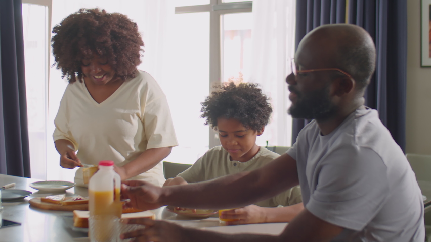 Cheerful African American woman making sandwiches for family breakfast while husband and little son eating and drinking juice at kitchen table Royalty-Free Stock Footage #1088793231