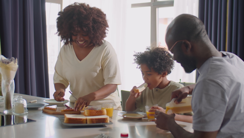 Cheerful African American woman making sandwiches for family breakfast while husband and little son eating and drinking juice at kitchen table | Shutterstock HD Video #1088793231