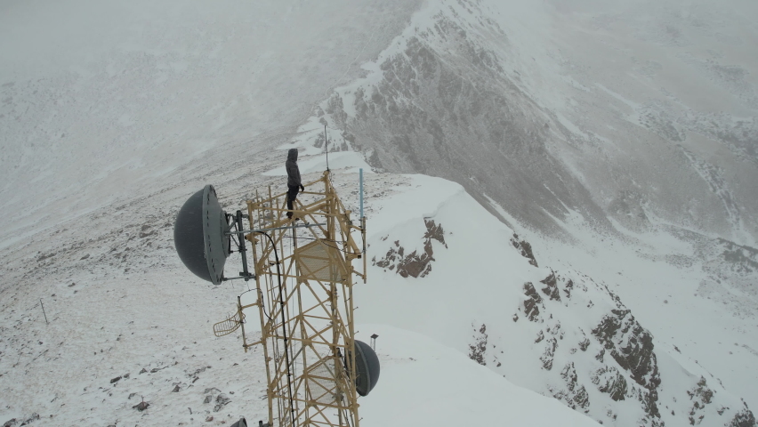 Aerial View of Man on Top of Communication Tower in Snow Capped Mountain Peak Raising Arms, Drone Shot