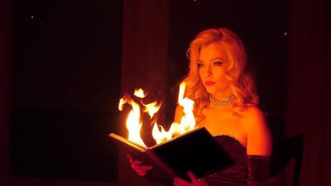 Glamor luxury lady in dress holds burning old book in her hands on dark background at night. Beautiful woman with magic fire book looking at camera. Mysterious ritual concept