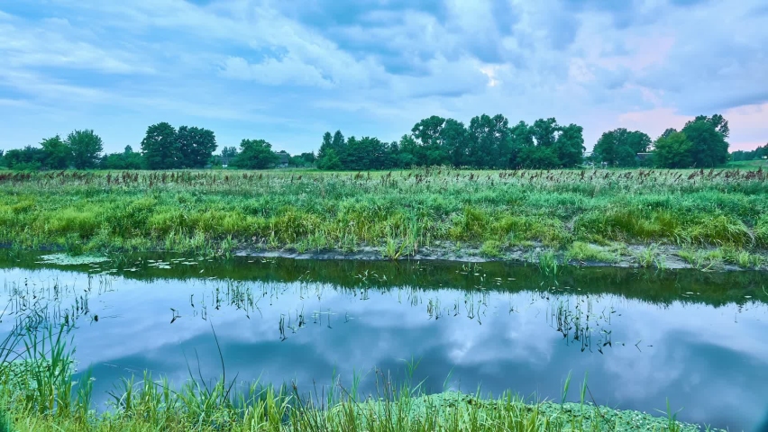 A small quiet river with overgrown banks. Summer countryside. Picturesque warm nature. In the distance is a village with wooden houses. Clouds float quickly across the sky.