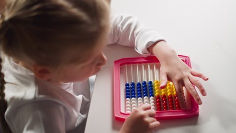 Cute little schoolgirl does sums moving plastic beads by left hand finger on colorful abacus at arithmetic lesson at home upper closeup slow motion