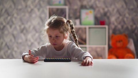 Active little girl with colorful toy abacus counts by finger and talks sitting at desk at arithmetic lesson against bookcase closeup slow motion