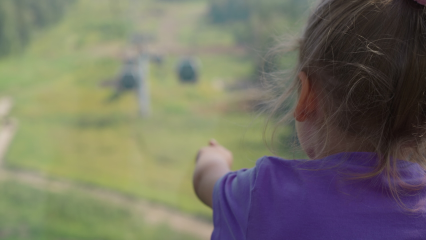 Blonde little girl with plait points to approaching cable car sitting at ropeway cabin window above forest clearing on sunny day close backside view slow motion