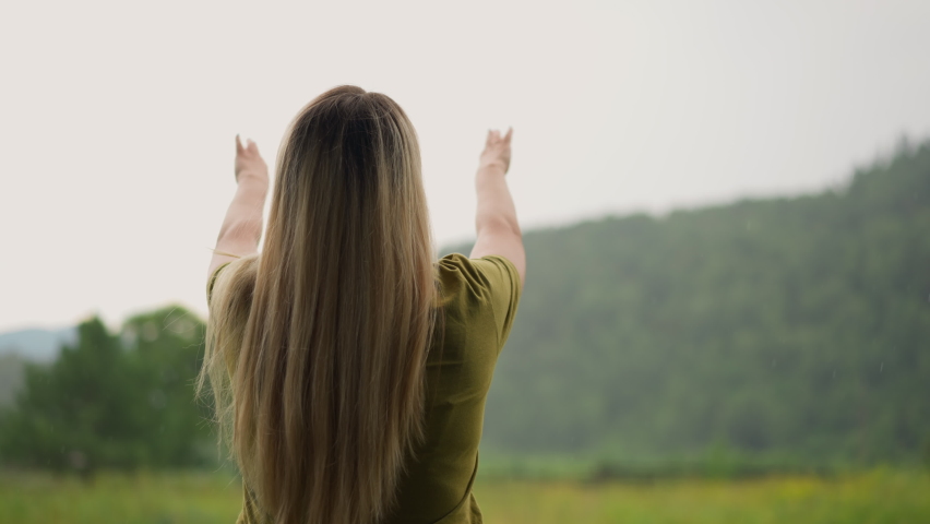 Cheerful young woman raise hands up happy of warm rain against green sloppy hill at eco resort on nasty day backside view slow motion