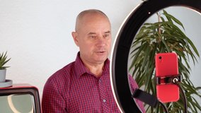middle-aged charming man, 60-65 years old blogger sits in front of ring light and red smartphone, emotionally talks about business, politics and records video, filmed on a 4K camera