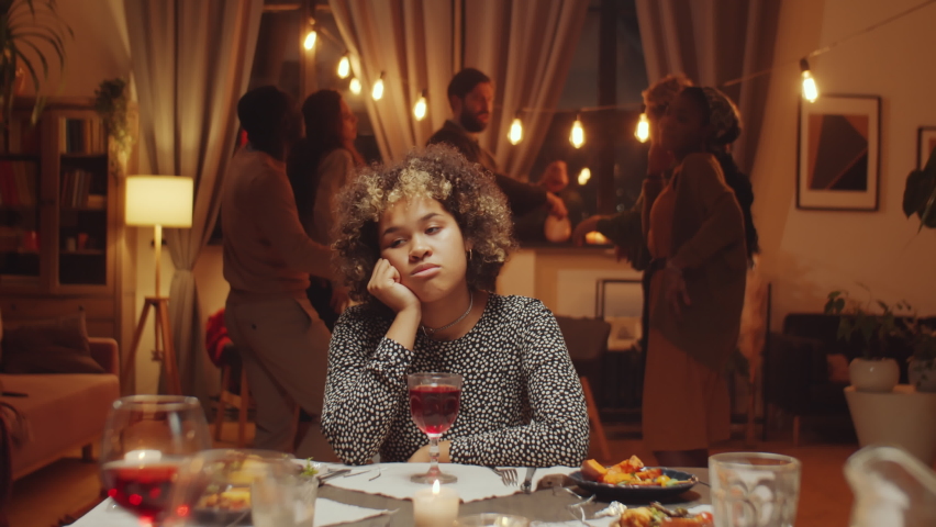 Slow motion of sad young Black woman sitting at dining table during birthday party, friends dancing behind her Royalty-Free Stock Footage #1088813779