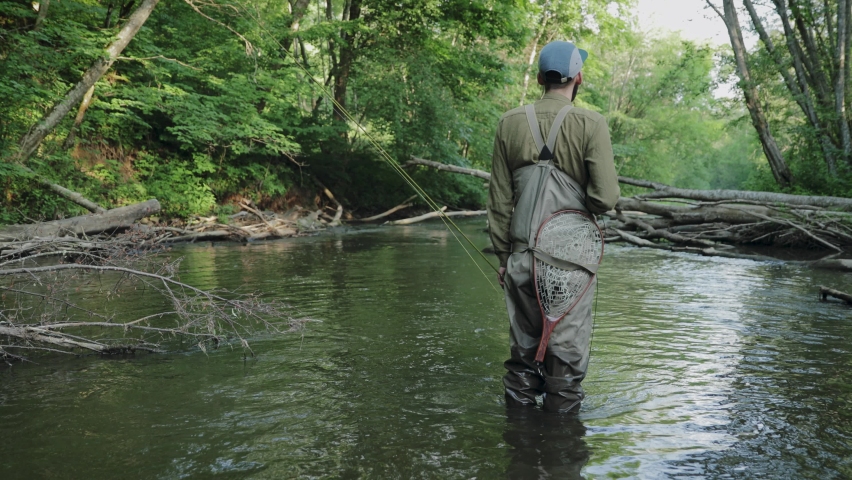fisherman standing in the middle of small wild fresh water mountain river. Person dressed in special outdoor activities clothing is looking around checking surroundings and searching for fishing spot.