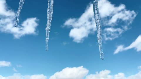 Three icicles hang and melt in sun on spring day. Blue sky and white clouds. The first warm days after cold winter. Icicles glisten in the sun. 4K footage.