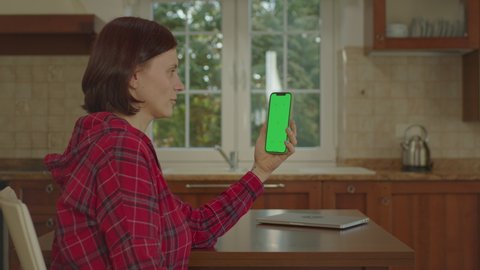 30s woman talking online to green screen holding mobile phone vertically sitting in the kitchen. Female laughing to vertical cell phone chroma key.