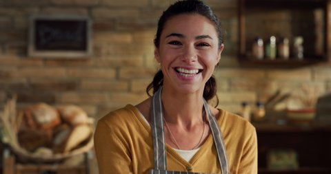 Cinematic close up shot of young female artisan baker is smiling in camera while kneading dough for preparation of pasta, pizza and other pastries in rustic bakery shop kitchen.