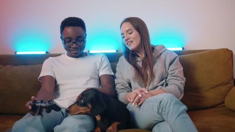 An African American young man and a Caucasian young girl are sitting on a sofa with a dog and playing a video game. Slow motion