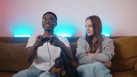 Dachshund lying on the couch looks in bewilderment as a young girl laughing take away a joystick from an african american guy. Slow motion