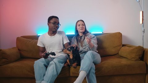 A cute dachshund is lying on a couch between a young couple who are playing video games and laughing. Slow motion