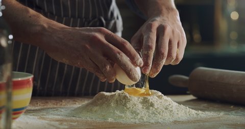 Cinematic close up shot of professional artisan baker chef is breaking egg in flour dough while preparing homemade pasta of fresh ingredients in rustic bakery kitchen.