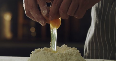 Cinematic close up shot of professional artisan baker chef is breaking egg in flour dough while preparing homemade pasta of fresh ingredients in rustic bakery kitchen.