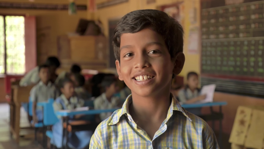 Close up shot of a cute smiling Indian Rural school boy in uniform looking at the camera with a group of kids or fellow student classmates sitting in classroom. concept of child education and literacy Royalty-Free Stock Footage #1088825181