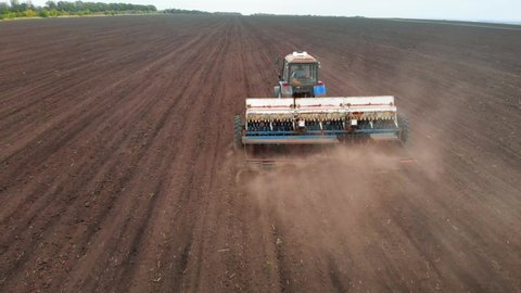 Drone sliding shot of a farmer in tractor seeding on sunny day, footage of sowing agricultural crops at field. Aerial view of a farmer in tractor seeding and cultivating land.