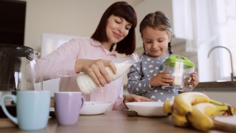Young beautiful mother pouring milk into daughters bows of cornflakes for breakfast in light modern kitchen. Little girl is waiting while her mom's hands pouring milk in a bowl for breakfast.