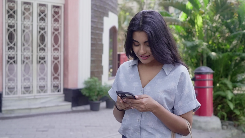 Cheerful Modern young urban Indian Asian beautiful Woman standing outdoors in a city using Mobile phone typing a message or texting with a smile on a face. Concept of fashion, trend, and technology Royalty-Free Stock Footage #1088826151