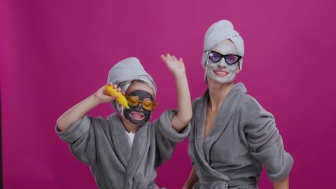 Funny mom and girl dancing in bathrobes having funny skincare party. Cute superstar daughter in golden sunglasses singing song performing on background.