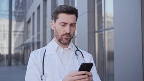 Male doctor physician standing near clinic outdoors holding mobile phone texting message turning head ahead, scrolling screen of smartphone sends online prescription to the patient during a pandemic.