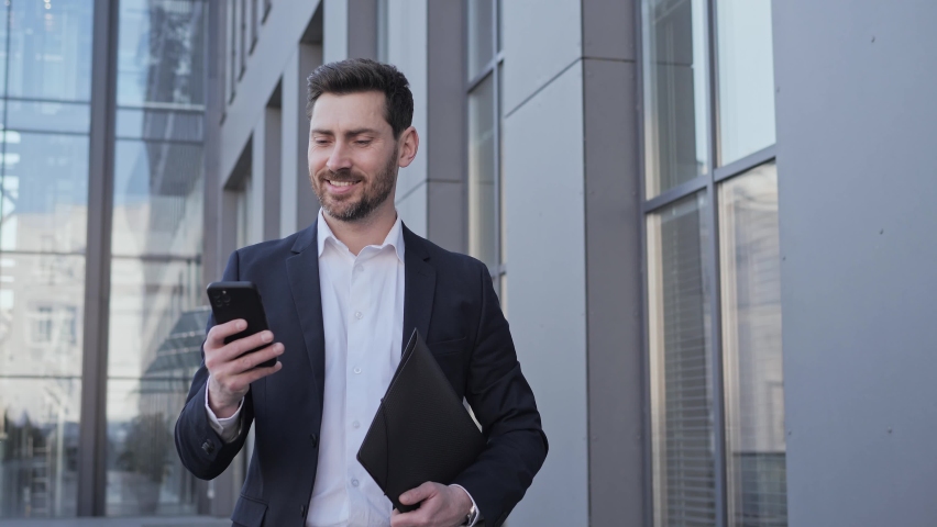 Handsome smiling man in stylish formal suit using smartphone app. Handsome businessman tapping scrolling browsing internet walking near office district holding folder with documents at city street.