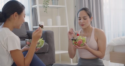 Two beautiful Asian woman eats  salad  from a bowl with sitting on the floor in the living room.Eat nutritious food.Yoga woman.Slow motion.Healthy lifestyle  concept.