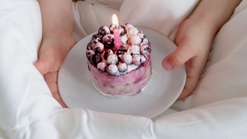 kid wrapped in white blanket is holding a mini birthday cake in hands with a pink candle. loving parents surprising his child with sweet dessert in bed. tasty and good looking mini cake with berries 