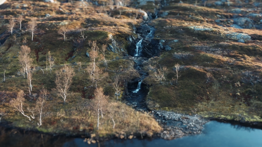 A mountain river cascades from the gentle slope. Moss, lichen, and withered grass cover the ground. Aerial vies, slow-motion, pan forward.