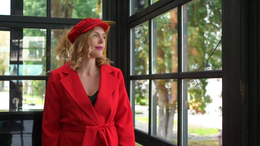 Elegant blonde woman in red suit and cap is waving her hand to somebody, spending time in the beautiful interior veranda