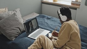 African American man in wireless headphones lying on bed at home, listening to female tutor and writing down some notes while having online lesson via video call on laptop