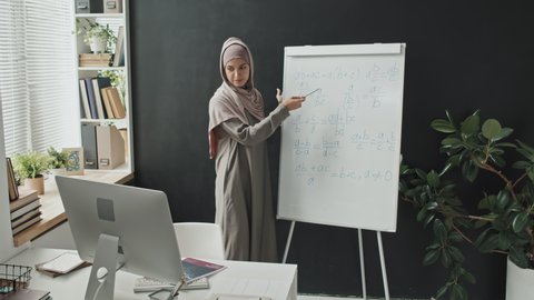 Muslim teacher in hijab explaining math formulas on flipchart while giving online lesson via video call on computer