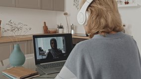 Senior woman in wireless headphones sitting at kitchen table and discussing business plan with Muslim female colleague via video call on laptop while working remotely during isolation