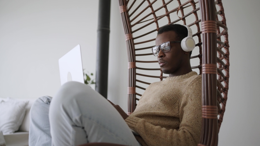Focused black male freelancer in glasses and headphones working on a laptop while sitting in a hanging rocking chair at home