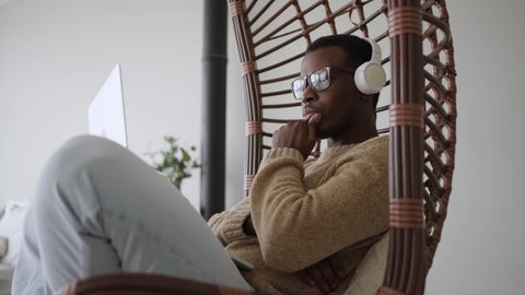 Focused black male freelancer in glasses and headphones working on a laptop while sitting in a hanging rocking chair at home