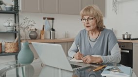 Senior Caucasian woman sitting at kitchen table, taking notes and talking via video call on laptop while taking online lesson at home
