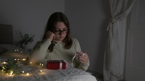 Upset and sad lonely girl sitting near the bed and touch white ribbon of gift in her bedroom with light garland and eucaliptus branch. Christmas o New Year alone, young triste lwoman in winter holiday