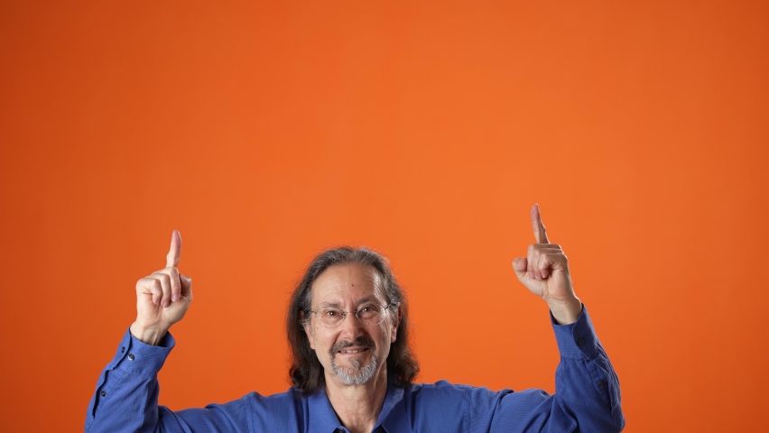 Happy promoter lively mature man 60s wears blue shirt pointing fingers up on workspace copy space mockup promo commercial area isolated on plain orange background studio portrait