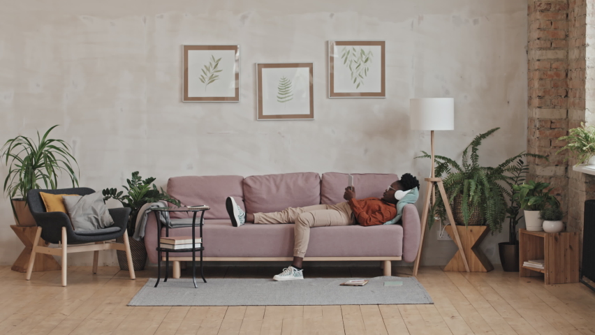 Wide shot of African American girl listening to music with wireless headphones while lying on sofa at home and using digital tablet